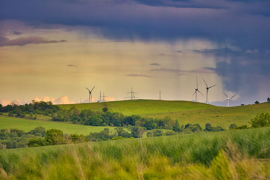 Dunkle Wolken mit Regenschwaden ziehen über einen Höhenrücken mit Grasland, Waldstreifen, Windenergieanlagen und Hochspannungsmasten.